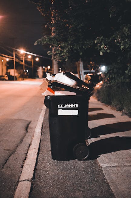 A black outdoor waste bin with the label 'St. John's' positioned on a paved sidewalk alongside a residential street at night. The bin is overflowing with a variety of rubbish, including cardboard boxes, plastic bags, and miscellaneous debris, some of which is leaning out or resting on top of the lid. The surrounding environment features dim street lighting, casting faint shadows on the pavement, and trees with dense foliage partially illuminated in the background. A row of streetlights and parked cars line the distant roadway, contributing to the quiet, urban nighttime scene. The concrete curb and the edge of the sidewalk frame the bin, indicating a typical domestic or community waste collection point. The scene reflects a scenario where private waste disposal or on-site rubbish accumulation may occur, aligning with the context of alternative rubbish removal services.