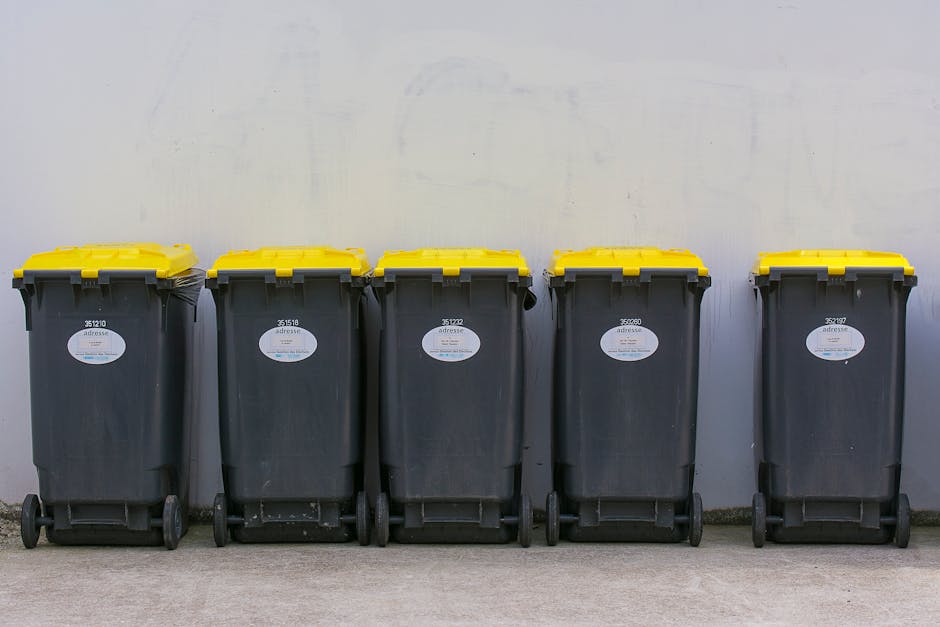 A row of five large, black wheeled rubbish bins with yellow hinged lids are lined up against a plain, light-colored wall on a concrete surface. Each bin has a white oval label with barcode and identification numbers. The bins are evenly spaced, positioned side by side, with the lids closed. The environment appears to be an outdoor area, possibly a driveway or designated waste collection zone, with natural lighting illuminating the scene without casting harsh shadows. The focus on these clearly identifiable waste containers suggests an independent or private waste collection service, such as those provided by Waste Collection Ruislip, supporting alternative methods of rubbish disposal apart from local authority services.
