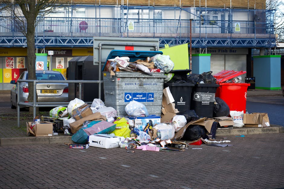 A waste collection worker wearing a high-visibility red and yellow vest stands at the rear of a large red rubbish truck, positioned on the side of a street. The truck's rear compartment is open, revealing mechanical lifting mechanisms and some waste inside. The worker appears to be managing or inspecting the waste, with a black plastic bag placed on the bumper. The scene is outdoors during daytime, with a cloudy sky and blurred background showing trees, power lines, and a small commercial building with signage. There is a small blue container on the ground near the truck, and a motorcycle parked next to it. The environment suggests an urban or suburban setting where private or independent rubbish collection services, such as those provided by Waste Collection Ruislip, are being carried out, with visible waste being prepared for disposal or collection.
